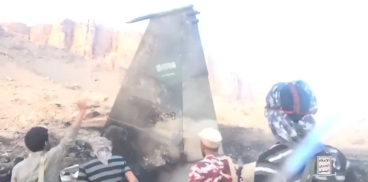 Yemeni fighters stand around the scorched wreckage of a large downed drone in a desert canyon, with a Houthi military-media watermark in the corner.